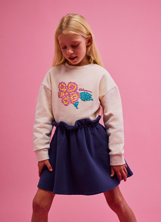 Schickes Outfit für junge Mädchen mit den neuen Kleidungsstücken von Vilebrequin: ein besticktes Sweatshirt mit Rundhalsausschnitt und Blume und ein marineblauer Baumwollminirock