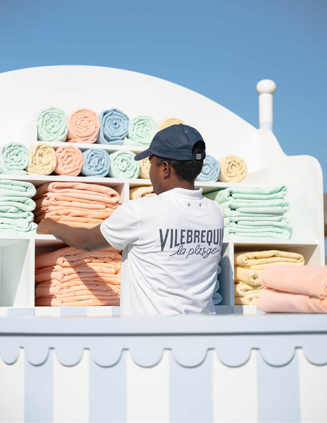 A beach attendant wearing a Vilebrequin shirt organizing rolled towels on white shelves.