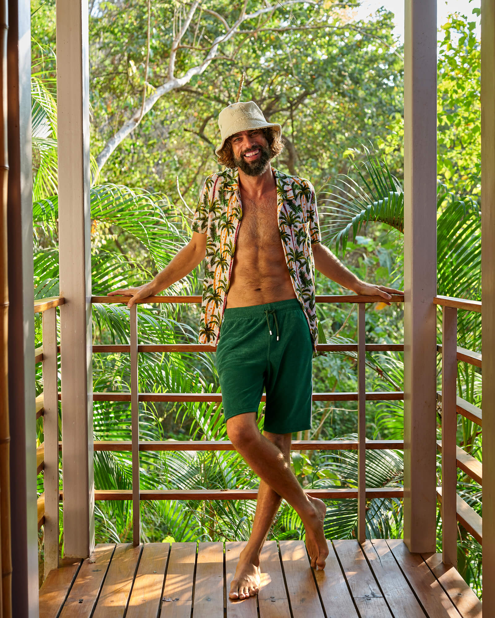 Colección de loungewear de verano para hombre Hombre sonriente con bermudas de loungewear verdes, camisa abierta con estampado de palmeras y gorro piluso beige en un balcón tropical