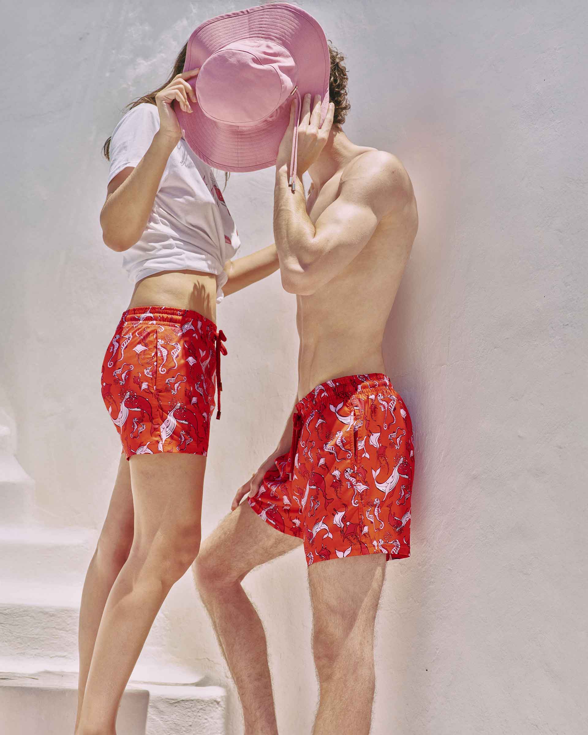Pareja con shorts de baño rojos a juego y estampado de ballenas blancas, escondiendo juguetonamente sus rostros detrás de un gran sombrero rosa.
