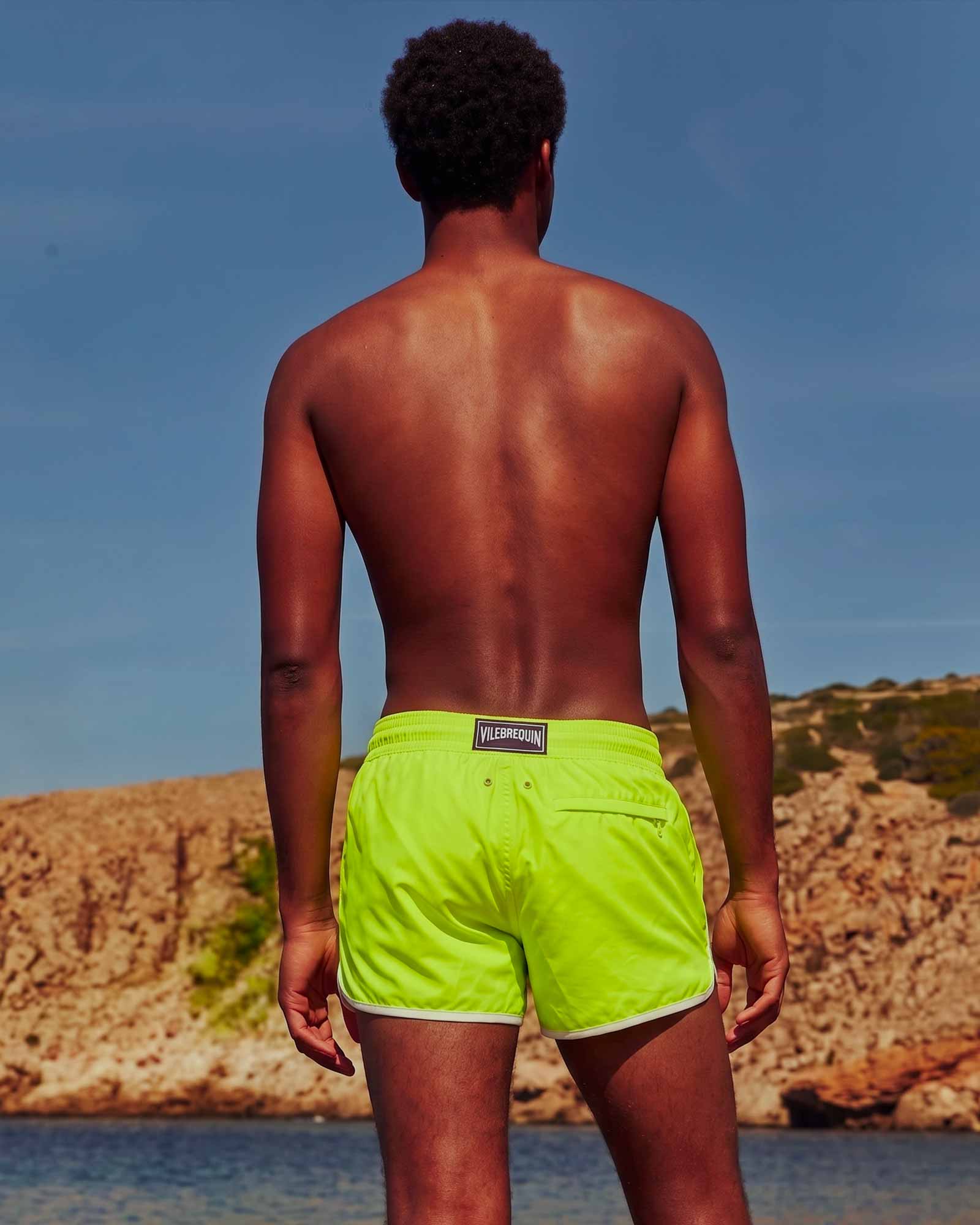 Costume da bagno giallo Vilebrequin Uomo di spalle con costume da bagno giallo fluo di fronte al mare.