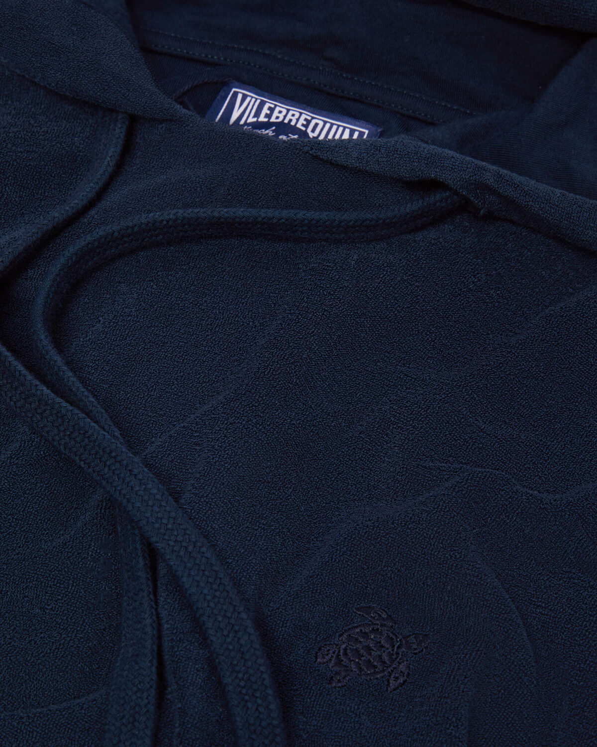 Navy blue terry cloth loungewear hoodie Close-up of a navy blue terry cloth loungewear hoodie featuring drawstrings, embroidered turtle logo, and Vilebrequin label